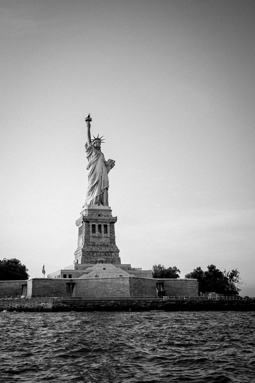 Black-and-white photo of the Statue of Liberty on Liberty Island shot from a sailboat on the Hudson River, NYC harbor.