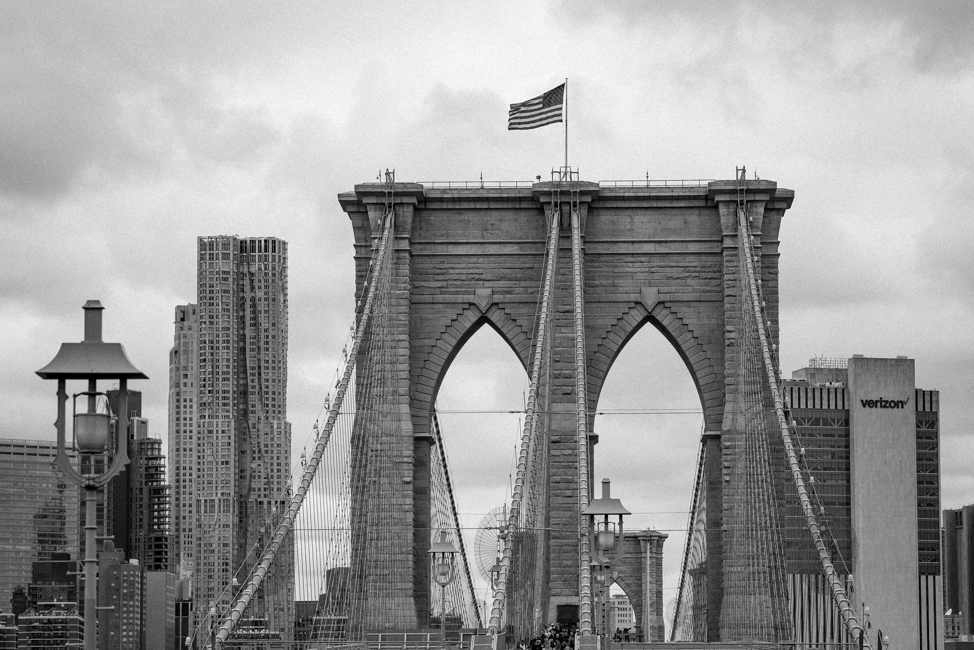 Black-and-white photo of the Brooklyn Bridge towers from a distance with the Lower Manhattan skyline and an American flag, NYC.