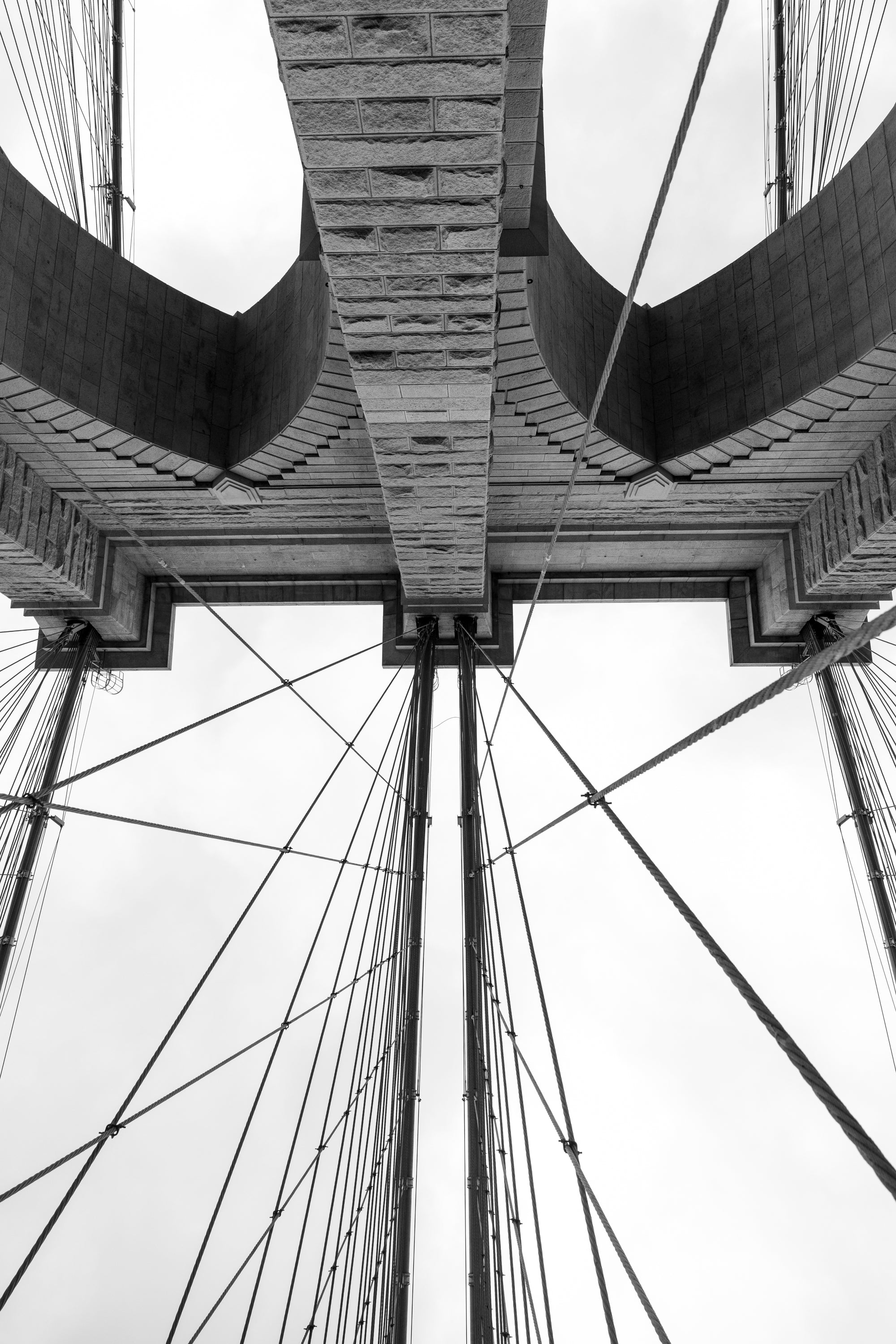 Black-and-white photo of the Brooklyn Bridge granite tower shot from directly underneath, gothic arches and radiating cables, NYC.