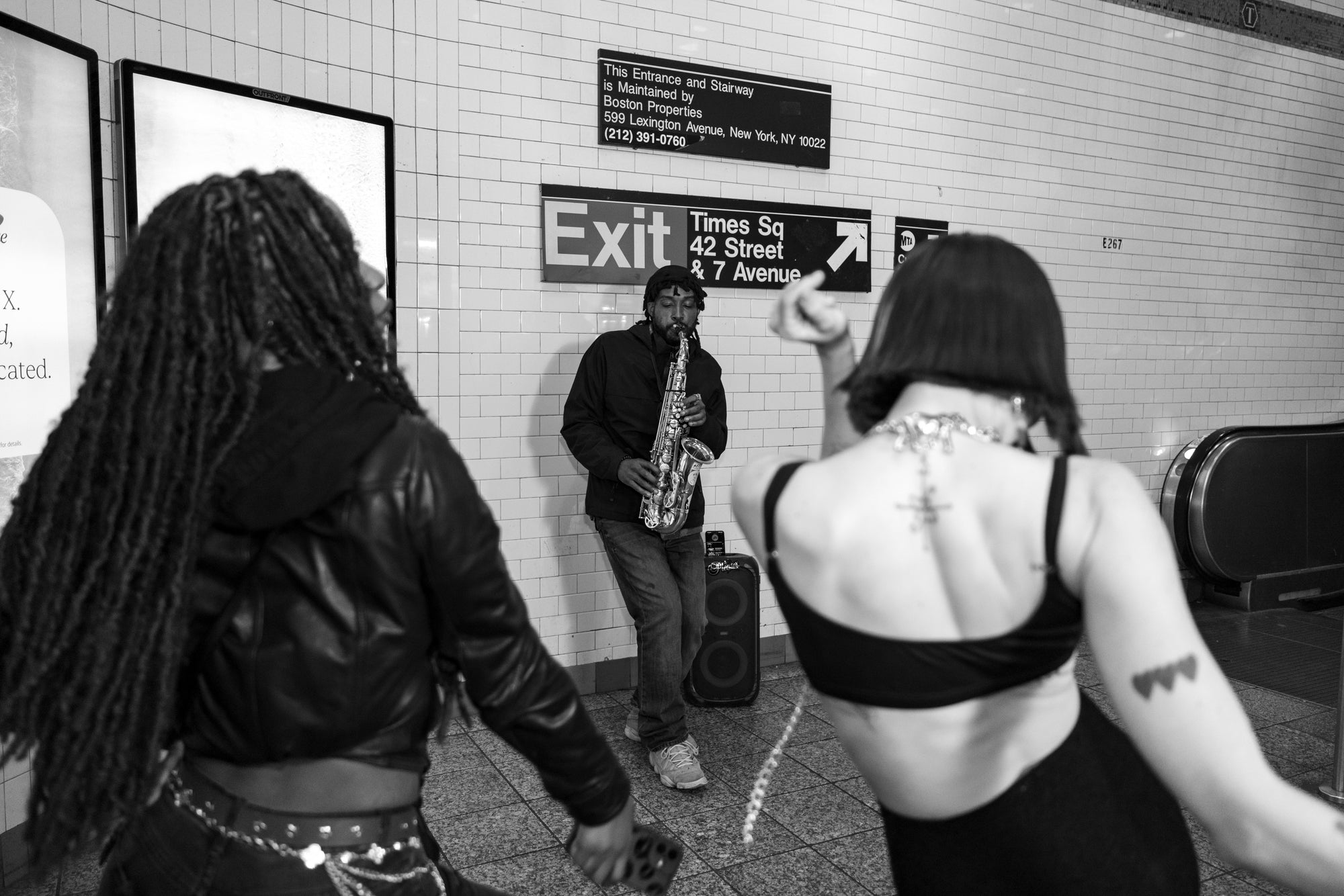 Black-and-white photo of a saxophone busker and two women dancing under the Times Square 42nd Street Exit sign, NYC subway.