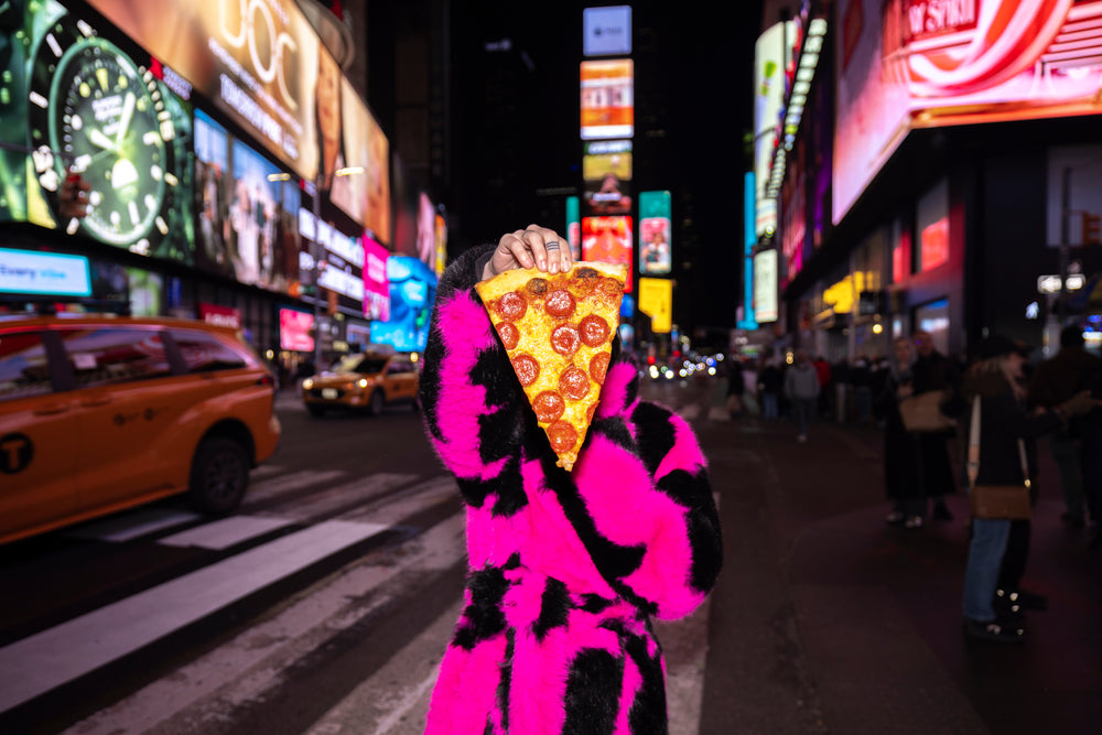 Color photo of a person in a pink-and-black fur coat holding a slice of pizza up to her face in Times Square at night, NYC.