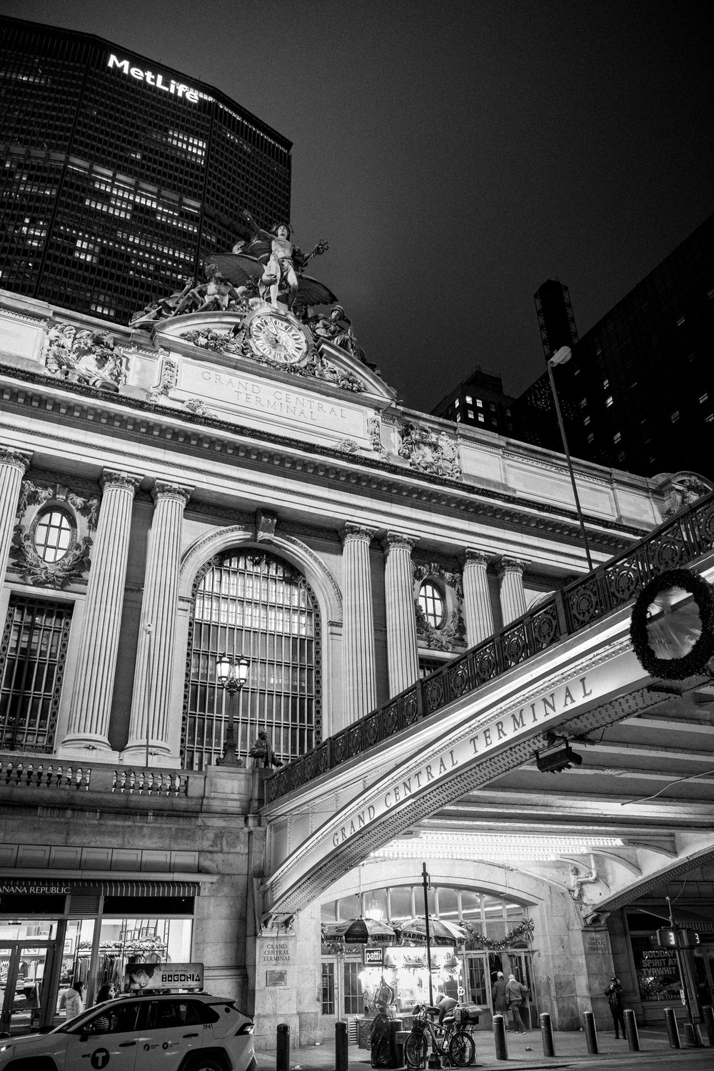 Black-and-white photograph of Grand Central Terminal at night from the left side of East 42nd Street with the Mercury statue, clock, and MetLife Building behind, NYC.