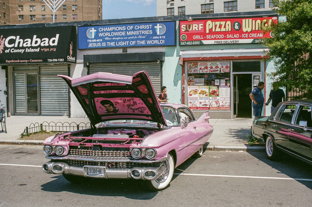 Color photo of a 1959 pink Cadillac with Elvis painted in the open trunk, parked in front of Chabad and Surf Pizza & Wings, Coney Island.