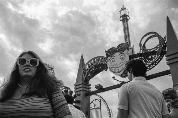 Black-and-white photo of a woman in oversized sunglasses in front of the Tillie face, Luna Park drop-tower, and a roller coaster at Coney Island, Brooklyn.