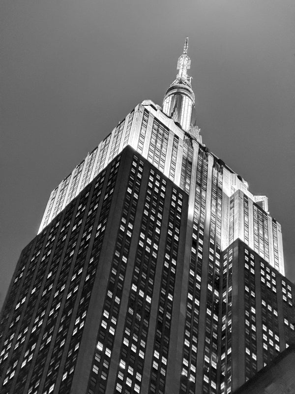 Black-and-white photograph of the Empire State Building shot from the sidewalk looking up, art deco crown and antenna, midtown Manhattan.