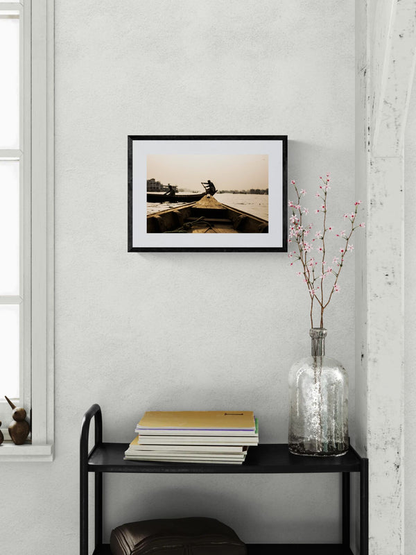 Dhaka Rower at Sunrise, Buriganga River framed in black on a gallery wall with a viewer in a straw hat in front — Good Beans NYC.