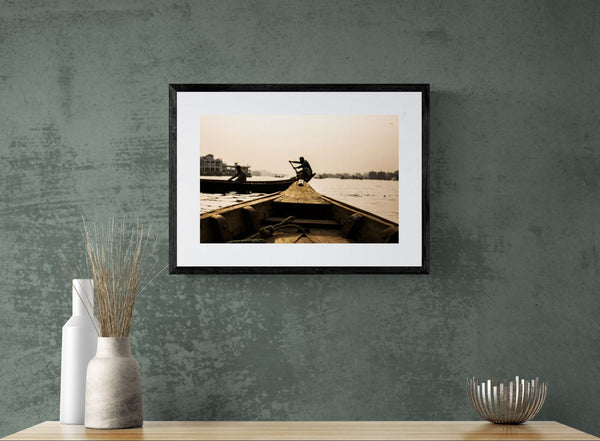 Dhaka Rower at Sunrise, Buriganga River framed in black above a beige sofa, sunlit yellow plaster wall — Good Beans NYC.