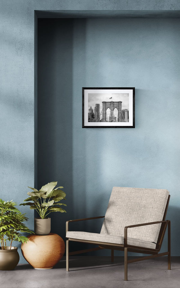 NYC Brooklyn Bridge from Afar, Manhattan Skyline framed in black above a beige sofa, sunlit yellow plaster wall — Good Beans NYC.